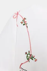 Camiseta blanca de manga corta y cuello redondo, con bordados de flores y lazos en tonos rosados y verdes. Incluye un bordado de un pájaro y texto en negro.