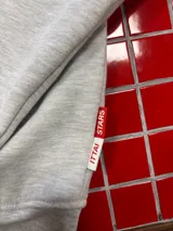 Buzo con capucha de color gris melange, con diseño estampado en la espalda que incluye el logo de la marca y elementos gráficos como un reloj y una botella. Presenta un corte holgado y puños elásticos.