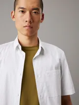 Camisa de manga corta Calvin Klein confeccionada en popelín de algodón elástico con diseño de rayas verticales. Presenta cuello en punta, cierre frontal de botones, bolsillo de parche en el pecho con logo bordado y bajo recto.