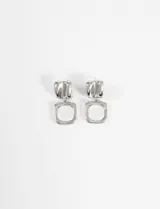 Caravanas colgantes de metal plateado, compuestas por una pieza superior de forma irregular y texturizada, y un dije inferior en forma de cuadrado hueco.