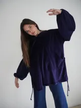 Buzo tipo canguro oversize de tela plush, color azul claro. Presenta capucha, cierre frontal parcial con cordón turquesa y bolsillos canguro frontales. Las mangas son largas y abullonadas con puños ajustados.