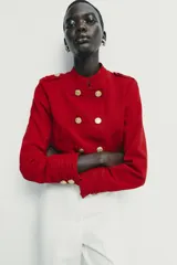 Cazadora de estilo militar en color rojo, con cuello redondo, manga larga y cierre frontal mediante alamares con botones dorados. Presenta trabillas con botón en los hombros y detalle de costuras en los puños.