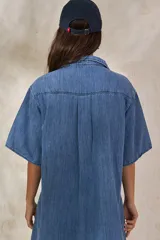 Vestido corto de denim azul, corte camisero holgado, mangas cortas amplias, cuello clásico, cierre frontal con botones, bolsillo aplicado en el pecho y pequeño bordado lateral en el dobladillo.