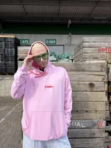 Canguro de tela deportiva color rosa, con capucha, cordones rojos y logo bordado en el pecho. Presenta un bolsillo frontal tipo canguro y diseño holgado.