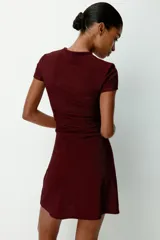 Vestido corto entallado de color bordó, con escote redondo y mangas cortas. Presenta un diseño con pliegues laterales en el hombro y la cintura que crean un efecto drapeado en el frente.