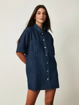 Vestido corto de denim azul oscuro, con cuello camisero, mangas cortas, cierre frontal de botones y dos bolsillos en el pecho.