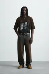 Camiseta de punto de algodón con corte boxy fit, cuello redondo y manga corta. Presenta un estampado frontal con efecto desgastado en tonos blanco y rojo sobre un fondo marrón oscuro/verde oliva lavado.