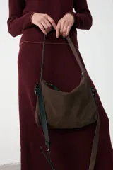 Cartera de hombro confeccionada en gamuza color chocolate, con cierre superior y doble sistema de correas: una fija de cuero liso negro para llevar al hombro y una correa larga ajustable y extraíble de gamuza y cuero negro para usar cruzada.