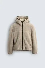 Cazadora de corte relajado confeccionada en tejido de textura borreguillo color beige. Presenta cuello subido con capucha ajustable, cierre frontal de cremallera en contraste verde, mangas largas y bolsillos laterales. El interior está forrado en tejido técnico.