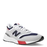 Championes urbanos New Balance modelo 997R, con capellada en gamuza gris y detalles en azul marino y rojo. Presentan el logo "N" en los laterales y entresuela blanca.