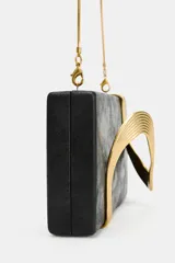 Bolso tipo clutch negro de formato rectangular con diseño rígido, adornado con un detalle metálico dorado de líneas onduladas en el frente. Cuenta con un cierre imantado y una correa de cadena metálica fina y dorada para llevar al hombro o cruzada.