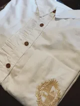 Camisa blanca de corte clásico con cuello camisero, cierre frontal de botones marrones y detalle de volado lateral. Presenta un bordado en hilo dorado en la parte inferior que representa el escudo nacional.