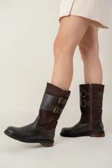 Bota de caña media confeccionada en una combinación de cuero liso y gamuza color chocolate. Presenta un diseño con cierre lateral, doble hebilla decorativa en el lateral externo y suela plana con pequeño tacón.
