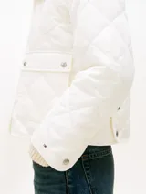 Campera acolchada Tommy Hilfiger de color blanco, confeccionada en poliéster reciclado con diseño matelasse en rombos. Presenta cuello camisero, cierre frontal con cremallera y botones a presión, dos bolsillos frontales de parche con solapa y detalle de logo bordado en el puño.