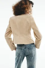 Blazer corto de cuero en color tostado, con cuello solapa, hombreras y manga larga. Presenta un diseño evasé en el bajo y un cierre frontal cruzado con dos botones.