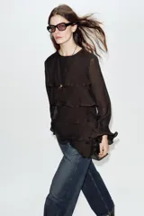 Blusa de cuello redondo y manga larga, color marrón oscuro. Presenta volantes en el frente y en los puños, y un bajo asimétrico. Cierre en la espalda con abertura y botón forrado.