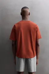 Camiseta de ajuste relajado (relaxed fit) color terracota, confeccionada en tejido técnico. Presenta cuello redondo, mangas ranglán cortas y un parche negro con logo en el bajo.