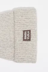 Gorro tipo beanie de punto texturizado en color gris claro con destellos plateados. Presenta un parche rectangular en la parte frontal con el logo de la marca en color marrón.