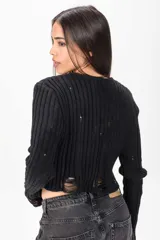 Cardigan corto de tejido de punto acanalado en color negro, con mangas largas y detalles desgastados o rotos en el frente y puños. Cierre frontal con pequeños botones redondos.