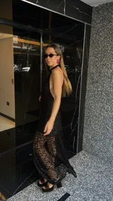 Vestido largo de encaje negro con cuello halter y espalda descubierta. Tiene un corte a la altura de la rodilla y un ruedo largo transparente.