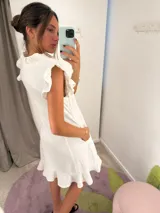 Vestido midi de color blanco con escote en V, cierre frontal abotonado y mangas cortas con volados. Presenta un diseño fluido con falda evasé y detalle de volado en el ruedo.