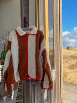 Campera de corderito con diseño de rayas verticales en colores marrón y crema, con cierre frontal y detalles de ribete en color rojo.