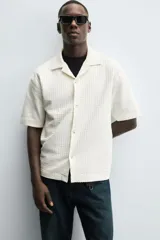 Camisa de manga corta con cuello bowling y calce relaxed fit, confeccionada en tejido seersucker elástico con textura. Presenta un diseño a rayas verticales en tonos oscuros y cierre frontal con botones.