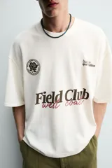 Remera de algodón color crudo, con textura de tejido abierto, cuello redondo y mangas cortas. Presenta estampas en contraste en el frente con los textos 'Field Club', 'West Coast', gráficos deportivos y un escudo circular.