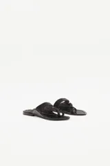 Sandalia plana de cuero negro con diseño de tiras trenzadas que cruzan el empeine y se sujetan entre los dedos. Presenta costuras decorativas en el borde de la plantilla y suela plana.