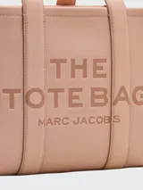 Cartera tipo tote de cuero granulado color tostado, con diseño estructurado y rectangular. Presenta doble asa superior fija y una correa larga ajustable y extraíble. En el frente, destaca el logo estampado con la inscripción 'THE TOTE BAG MARC JACOBS'.