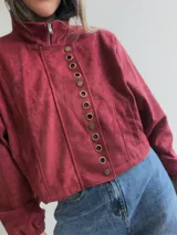 Chaqueta corta de efecto gamuza en color azul marino, con cierre frontal y solapa decorada con una hilera de aros metálicos. Presenta mangas tipo globo, ajuste lateral en la cintura y forro interior.