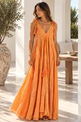Vestido largo color dorado, confeccionado en tela semitransparente con diseño bordado floral y detalles de brillo. Presenta escote en V, tirantes finos ajustables con borlas en los hombros, y corte amplio y fluido con gran movimiento.