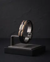 Pulsera masculina con diseño de placa central en acero con baño de oro rosa y detalles en negro, montada sobre una correa de cuerina negra con textura.