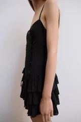 Vestido corto negro de tirantes finos ajustables y escote en V. Presenta un diseño ajustado con drapeado central y cordones cruzados laterales tipo corsé, y falda corta con tres capas de volantes escalonados.
