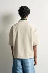 Sobrecamisa boxy fit de algodón color beige, de manga corta, con cuello solapa, cierre frontal de botones y aplicación de texto en parches rosas a contraste en el delantero.