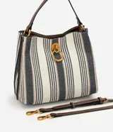 Cartera bucket pequeña de Satinato con diseño de rayas verticales en tonos blanco, azul marino y beige, confeccionada en tejido estructurado. Cuenta con asa de mano fija, asa larga desmontable de cuero sintético marrón, detalles de herrajes metálicos dorados, argolla metálica colgante en el frente y cierre superior.