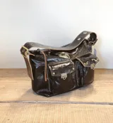 Bolso de hombro confeccionado en cuero con acabado charolado brillante. Presenta dos bolsillos frontales con solapa y cierres metálicos, correa ancha con ojales decorativos y herrajes en tono bronce.