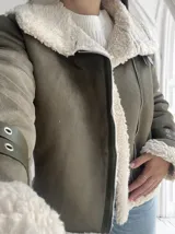 Chaqueta verde militar de gamuza sintética con interior de piel sintética color crema. Tiene cierre frontal con cremallera, cuello con solapas grandes y cinturón con hebilla en los puños.