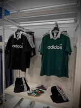 Remera polo Adidas Originals JSY negra con cuello blanco en V, tres rayas blancas en las mangas y logo Trefoil bordado. Confeccionada en tejido liviano y transpirable, con un corte regular.