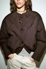 Cazadora corta acolchada de color negro, con cuello redondo, manga larga y cierre frontal con botones. Presenta un diseño de costuras geométricas y bolsillos laterales de vivo.