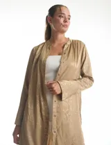 Túnica satinada color dorado con estampado animal print, de silueta oversize y fluida, largo maxi. Cuello a la base con abertura central y botones a tono. Manga larga de corte recto con puños amplios y cierre de botones. Espalda con canesú horizontal recto del cual nace un tablón central profundo. Bajo asimétrico redondeado con tajos laterales profundos, el largo de espalda mayor al largo delantero.