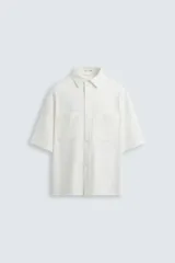 Sobrecamisa blanca de tejido con elasticidad, cuello solapa y manga corta. Tiene dos bolsillos de plastrón en el pecho y cierre frontal con botones a presión.