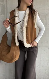 Chaleco de gamuza color camel con interior de corderito sintético, diseño sin mangas, borde frontal con ribete de corderito y detalle de tachas metálicas decorativas.