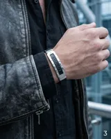 Pulsera de diseño moderno con estructura central de acero inoxidable y laterales de goma negra. Presenta un detalle central con el patrón de guarda griega grabado sobre fondo negro.