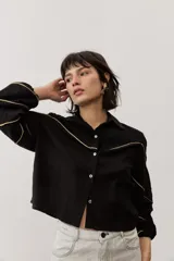 Camisa de manga larga color negro, con cuello camisero, cierre frontal con botones y detalles de ribetes contrastantes en color dorado. Presenta paneles superiores con textura y mangas abullonadas.