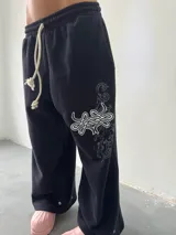 Pantalón de jogging negro de corte baggy, confeccionado en algodón. Presenta una estampa plastisol con bordado decorativo en la pierna izquierda, cintura elástica con cordón largo ajustable y botones en el ruedo para regular la botamanga.