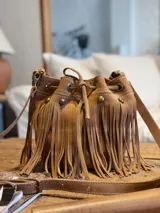 Cartera tipo bucket bag de gamuza color marrón, con diseño de flecos largos en todo el cuerpo, detalles de tachas metálicas y cierre mediante cordón ajustable.