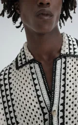 Camisa de manga corta color beige, tejida con punto abierto (símil crochet) y detalles en negro. Tiene cuello solapa, cierre frontal con botones y calce oversize.