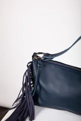 Cartera bandolera de cuero negro con flecos colgantes en el lateral, cierre superior y correa ajustable con argollas metálicas.