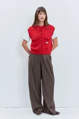 Chaleco de punto rojo con cierre frontal de botones, cuello redondo y dos bolsillos frontales con solapa. Presenta un diseño de tejido acanalado en los bordes y un pequeño parche decorativo en el pecho.